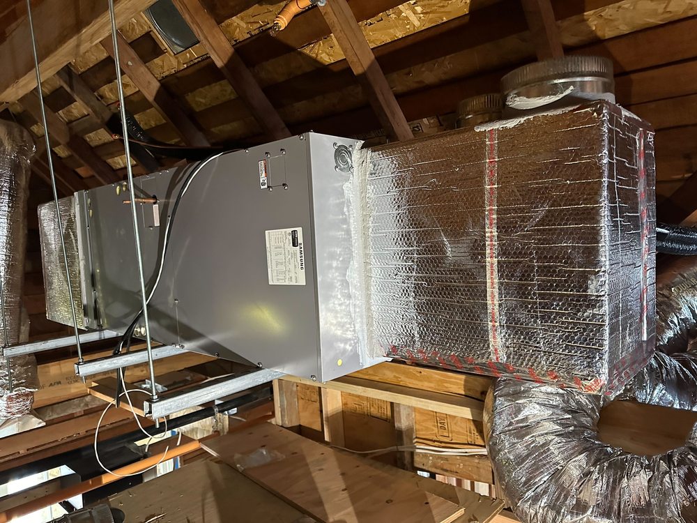 An air handler hung from the rafters in an attic, with foil-wrapped ductwork and a flex duct.
