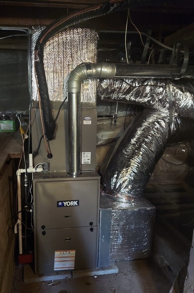 A new York gas furnace installed in a crawlspace, with insulated flex ducts and a flue pipe running up from it.